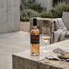 A bottle of wine with a black label and a partially filled wine glass on a stone table in an outdoor patio setting.