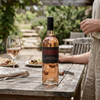 A bottle of Rosé wine from Luria Estate stands on a rustic wooden table in an outdoor patio setting, with a wine glass, plates, and part of a person visible in the background.